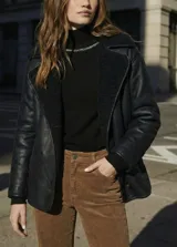 Chaqueta tipo gamulán de color negro con forro de corderito sintético al tono. Presenta solapas anchas, cierre frontal con botones y un diseño entallado al cuerpo.