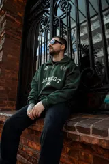 Buzo verde oscuro de algodón con cierre medio frontal, cuello alto y corte oversize. Estampado frontal con la frase "Against All" en color crema y detalles bordados.