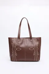 Cartera tipo tote bag en símil cuero color marrón oscuro, con bordados decorativos en blanco que evocan un estilo western. Cuenta con asas largas para llevar al hombro.