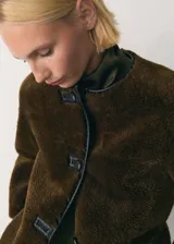 Chaqueta beige de poliéster con textura de borreguito, cuello redondo, cierre frontal con botones tipo trenca, ribetes en contraste de color negro, dos bolsillos delanteros y forro interior.