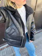 Campera negra estilo aviador, con cuello y forro de piel sintética. Cierre frontal con cremallera y hebilla en el cuello. Bolsillos laterales con solapa y puños ajustables con hebilla.