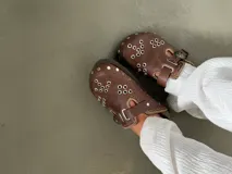 Zuecos marrones de cuero con hebilla metálica y detalles de bordado artesanal, tachas y remaches.
