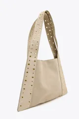 Bolso de hombro color beige, confeccionado en serraje, con tachas metálicas doradas en el cuerpo y las asas. Cuenta con un bolsillo interior y cierre mediante botón imantado.