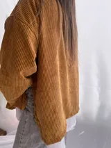 Buzo de pana fina color verde oliva, con diseño de corte holgado y mangas largas. Presenta una textura de cordón vertical característica de la pana y un pequeño parche decorativo en la parte superior de la espalda.