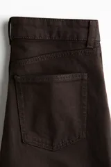 Jean de cinco bolsillos, tiro alto, color marrón oscuro o negro, con piernas anchas y rectas. Presenta cierre frontal con botón y remaches metálicos dorados.