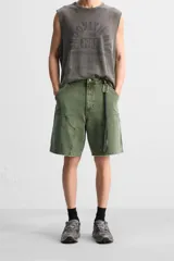 Bermuda color verde militar, de corte relaxed fit, con bolsillos frontales y traseros tipo plaqué, detalle de tira y bolsillo multifunción en la pierna, y bajo irregular.