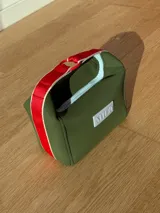 Neceser de neopreno color verde militar con cierre superior celeste, manija de agarre con detalle de cinta roja y logo de la marca en color gris. Posee forro y compartimento interno.