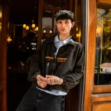 Chaqueta de jean negro con efecto lavado, corte recto, cuello clásico, cierre frontal con cremallera, bolsillos laterales y puños elásticos. Estampado con el texto "VENEFO COMPANY" en color naranja.