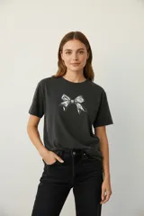 Remera de algodón color gris oscuro con cuello redondo y manga corta. Presenta un estampado frontal de un moño con textura de serpiente en blanco y negro, delineado con pequeños apliques de strass.