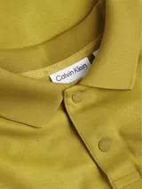 Chomba color ocre de corte recto, con cuello clásico y tapeta de dos botones. Presenta una banda horizontal en el pecho con el logo de Calvin Klein bordado en relieve.