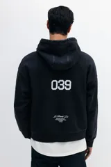 Sudadera tipo canguro de algodón pesado con efecto lavado oscuro, corte oversized y hombros caídos. Presenta estampado frontal en blanco con texto cursivo "Le Strade Di MMXXVI", dos banderas a cuadros, texto pequeño en la parte superior derecha y texto vertical en la manga izquierda. La manga derecha tiene un patrón de estrellas delineadas.