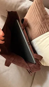 Bolso tote grande de material similar a gamuza color marrón oscuro, con dos asas de hombro y detalles de tachas plateadas a lo largo de los bordes verticales. Cierre de broche en la parte superior central.