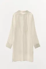 Camisa color crudo de corte holgado, confeccionada en hilatura con lino y tejido con efecto arrugado. Tiene cuello solapa, mangas largas con puños, bajo asimétrico con aberturas laterales y cierre frontal con botones.