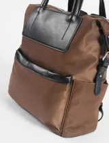 Mochila marrón de tela con detalles en cuero vegano negro, con doble asa de mano y correa de hombro ajustable.