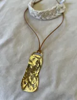 Collar con dije irregular de bronce con textura irregular y detalles grabados, colgando de un cordón de cuero sintético marrón. Incluye un choker de cuero sintético marrón.