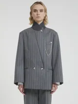 Blazer cruzado de lana gris con finas rayas blancas verticales (pinstripe). Presenta solapas anchas, doble botonadura con botones claros y un accesorio decorativo de cadena de perlas colgante prendido en la solapa izquierda.