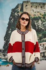 Cardigan de punto grueso, estilo universitario, con bloques de color azul marino, blanco y detalles en rosado y rojo en el cuello y bordes frontales. Presenta botones marrones grandes y el logo 'anthea EST.2016' bordado en el pecho.