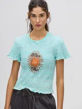 Remera corta de tela fruncida a rayas horizontales color celeste y blanco. Tiene cuello redondo, mangas cortas con volados y un estampado de un sol con texto en el centro.
