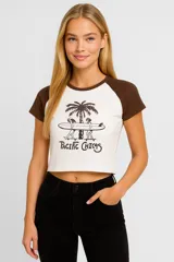 Remera corta color crema con mangas marrones tipo ranglán, cuello redondo marrón y estampado frontal con palmeras y texto "Pacific Chicas".