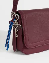 Bandolera color crudo de cuerina con solapa y correa regulable con hebilla. Incluye charm decorativo lateral y cierre con broche a presión.