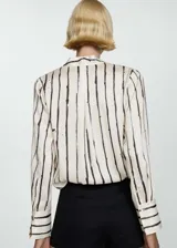Blusa de poliéster color blanco con estampado de rayas verticales negras, escote cruzado en V y mangas largas con puños abotonados.