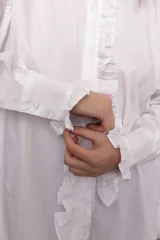 Camisa blanca de algodón con cuello clásico, cierre frontal con botones, mangas largas con puños abotonados y volados en el frente y los puños.