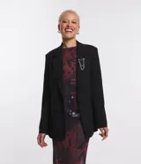 Blazer negro de sastrería con corte alargado, cuello con solapa, manga larga, botón delantero, bolsillos fake y cadena decorativa en la solapa.