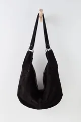 Bolso tipo hobo de gamuza negra, con forma holgada y cierre principal de cremallera. Se lleva al hombro con dos asas de correa delgadas sujetas con argollas metálicas plateadas.