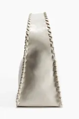 Cartera de mano color crudo, con forma rectangular y asa corta. Presenta detalle de tira entrelazada en los bordes y cierre superior.