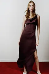 Vestido midi de tejido satinado color marrón oscuro (ciruela/vino), con escote drapeado (cowl neck) y tirantes finos. Presenta un panel tipo corsé con varillas y corchetes en la cintura, y una abertura lateral en la falda.