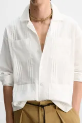 Camisa blanca de corte relaxed fit confeccionada en tejido de algodón y lino. Tiene cuello bowling, manga larga con puños abotonados, jaretas en el delantero, bolsillos de parche en pecho y cadera, bajo con aberturas laterales y cierre frontal con botones.