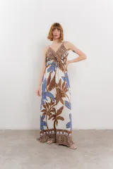 Vestido largo con estampado de hojas en tonos verdes y marrones sobre fondo blanco. Tiene breteles finos, escote en V y detalle de lazo en el frente. La falda es amplia y con vuelo.