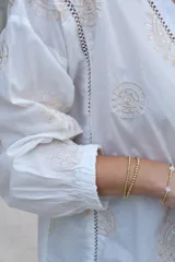 Blusa blanca de manga larga con elástico en puños, escote en pico y cierre frontal con botones. Detalles de bordados con hilado en tonalidad camel y acabado con entredoses a tono.