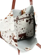 Bolso tote de cuero con pelo, estampado de vaca en blanco y marrón. Tiene asas de cuero marrón y un pequeño parche de cuero marrón con logo grabado en el frente.