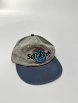Gorro tipo gorra de béisbol, color gris claro con visera azul. Presenta un bordado frontal con el texto "Soft Rock Cafe" y un diseño de un globo terráqueo con detalles en rojo, azul y verde.