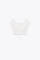 Top crop blanco de escote cuadrado y manga corta abullonada. Detalle de lazada en el pecho y volantes en el escote y el bajo.