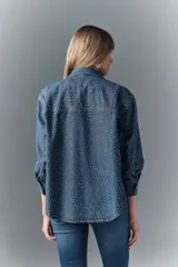 Camisa de denim azul con apliques metálicos plateados, de calce amplio, cuello camisero, cierre frontal con botones a presión, mangas largas con puños abotonados y dos bolsillos con solapa abotonada en el pecho.