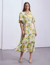 Vestido largo camisero con estampado floral amarillo y verde sobre fondo blanco. Tiene cuello camisero, mangas cortas con volados, cintura con lazo ajustable y falda escalonada con volados.