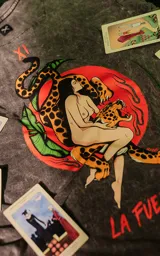 Remera gris con efecto nevado, de corte oversize y manga corta. Estampado con una mujer desnuda abrazando la cabeza de un jaguar, rodeada de hojas y con el texto "La Fuerza" en rojo.