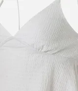 Blusa corta blanca de poliéster y elastano, con textura arrugada, escote en V, breteles finos y bajo asimétrico con volados laterales.