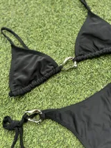 Conjunto de bikini negro con corpiño triangular con argolla metálica y bombacha colaless con tiras para atar y argollas en los costados.