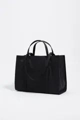 Bolso shopper de nylon negro, estructura blanda, con doble asa de mano y cierre principal de cremallera. Presenta una etiqueta logotipada en el frente y herrajes metálicos lacados.