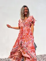 Vestido largo de corte evasé con estampado floral en tonos rosados y anaranjados. Tiene escote en V cruzado, mangas cortas con volantes y falda con volantes asimétricos.