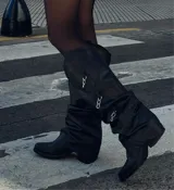 Bota alta de cuero negro con caña arrugada y detalles de cadenas colgantes.