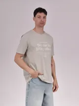 Remera color beige con estampado frontal con la frase "We rise by lifting others" y otros textos en tipografía pequeña.