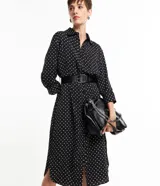 Vestido midi tipo chemise de tela satinada negra con lunares blancos, cuello solapa, abotonado delantero, mangas largas y cinturón con hebilla.