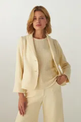 Chaqueta color crema de corte recto con cuello, solapa y bolsillos ojal. Forro solo en las mangas y terminaciones envivadas. Esquinas redondeadas y mangas con corte en puño que permiten transformarla en manga 3/4. Más larga en la espalda que en la delantera. Abrochadura con dos botones.