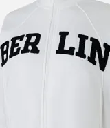 Chaqueta deportiva blanca con cierre frontal, cuello alto y puños ajustados. Presenta la inscripción "BERLIN" en letras negras y gruesas en el pecho.
