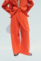 Pantalón de vestir color naranja, de tiro alto y pernera ancha. Presenta doble pliegue frontal, trabillas en la cintura, bolsillos laterales y falsos bolsillos de vivo en la espalda.
