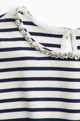 Remera de punto acanalado a rayas horizontales blancas y azul marino, con cuello redondo adornado con strass brillante. Presenta abertura de cerradura con botón en la nuca y bajo recto. Corte ajustado.